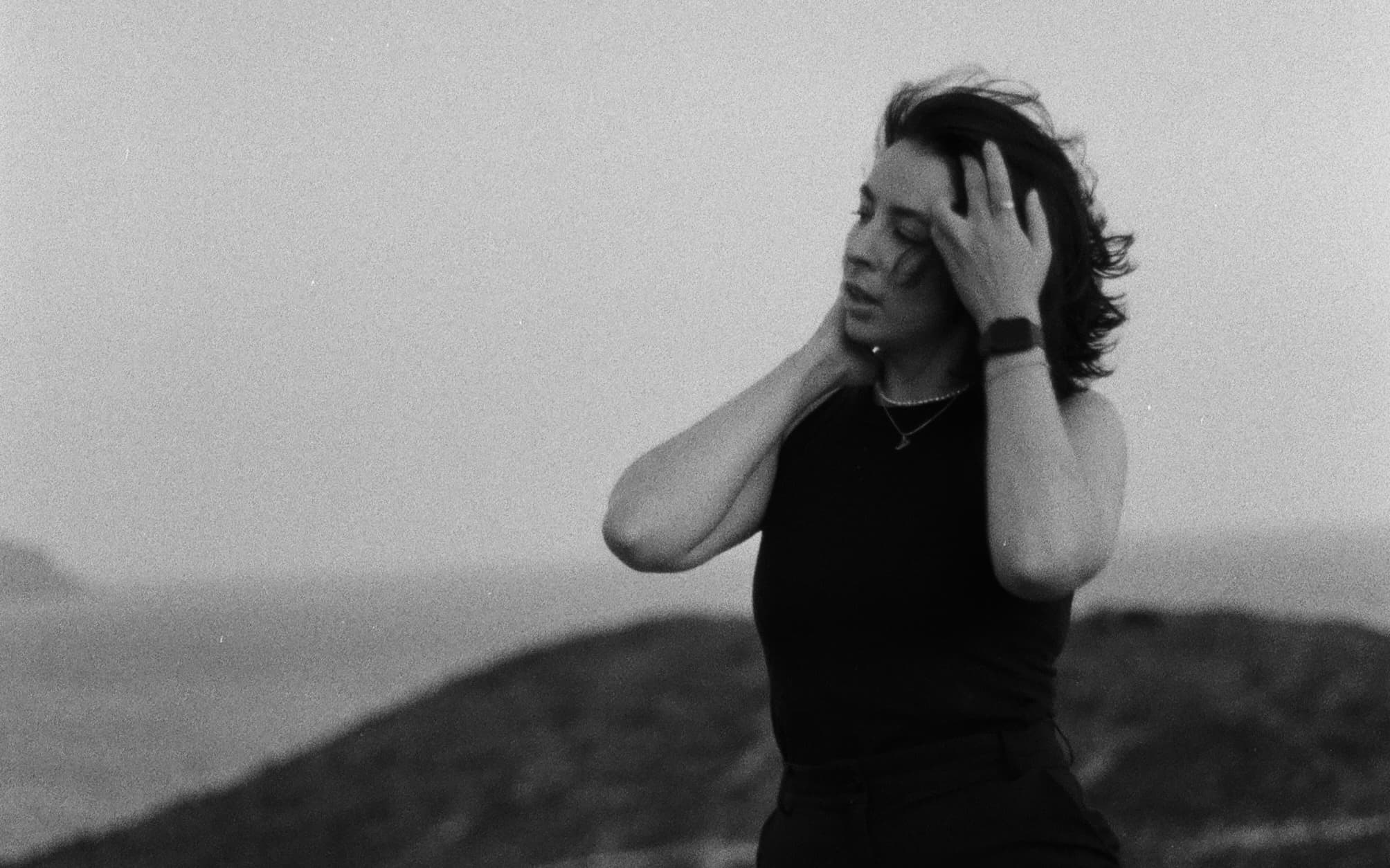 Black and white image of a woman standing outdoors, eyes closed, hands on her head, with a blurred landscape in the background.
