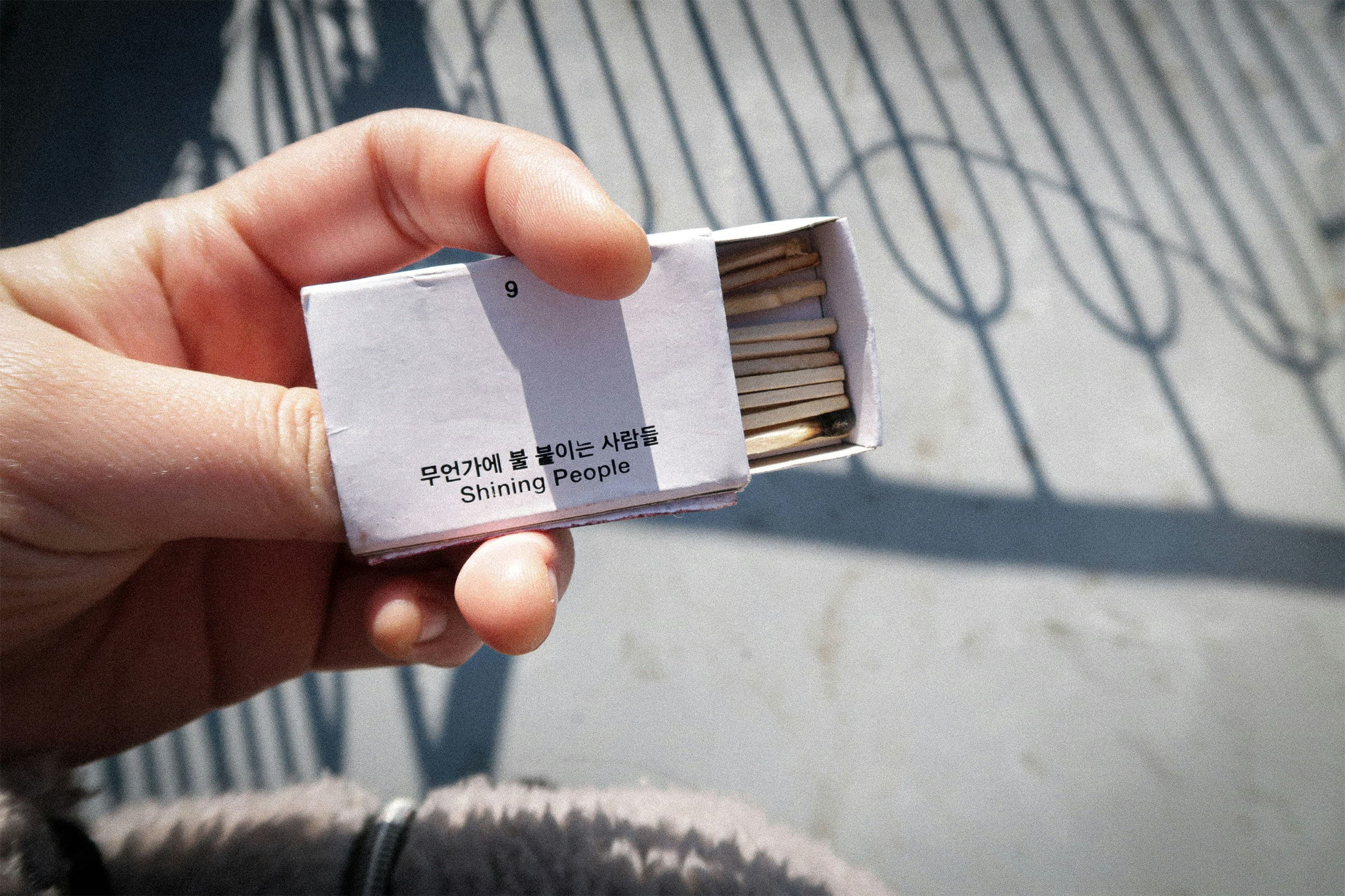 Hand holding an open box of matches labeled "Shining People" with Korean text, against a sunny outdoor background with shadows.
