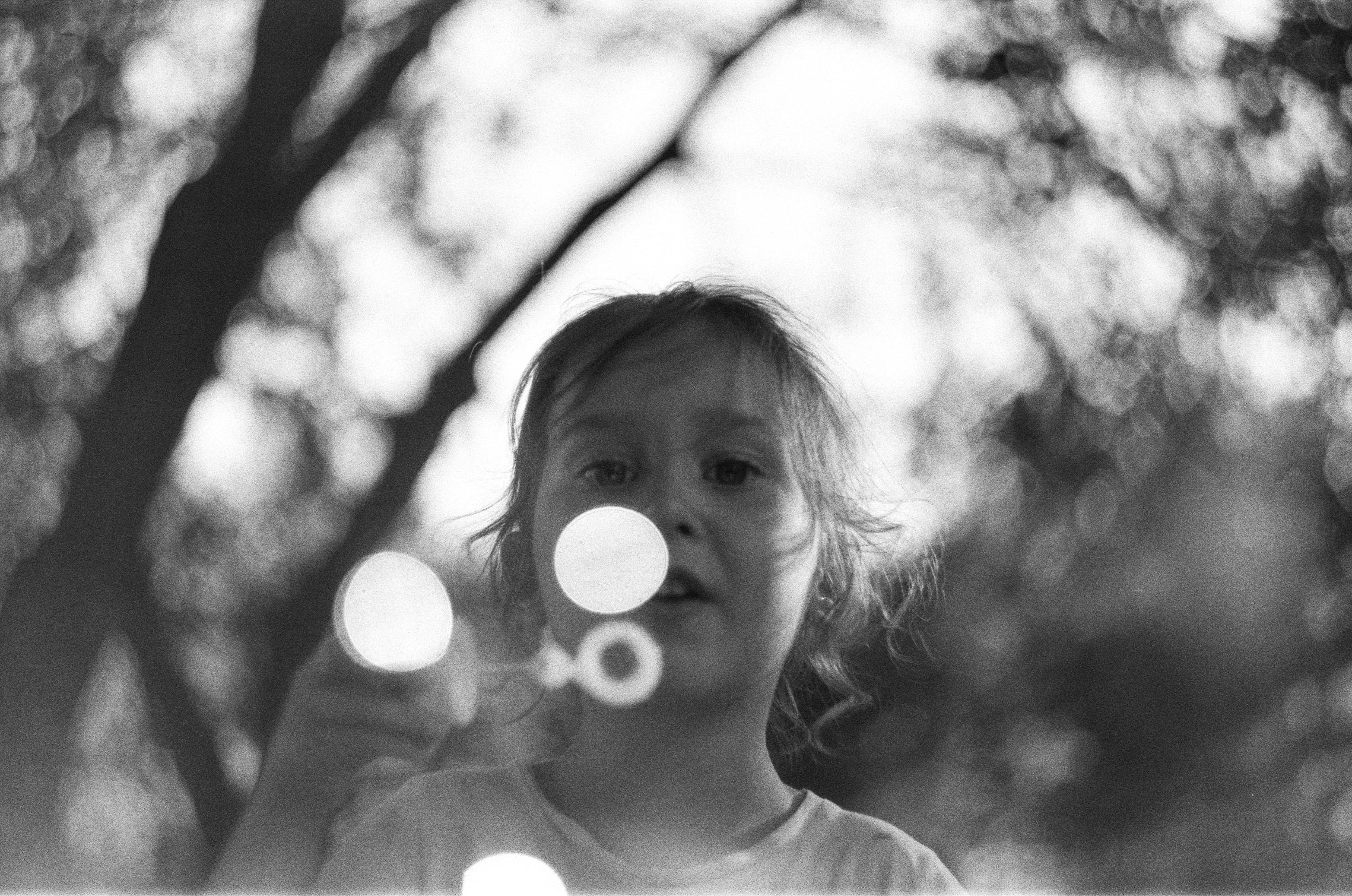 Black and white photo of a child blowing bubbles outdoors, with blurred trees in the background.