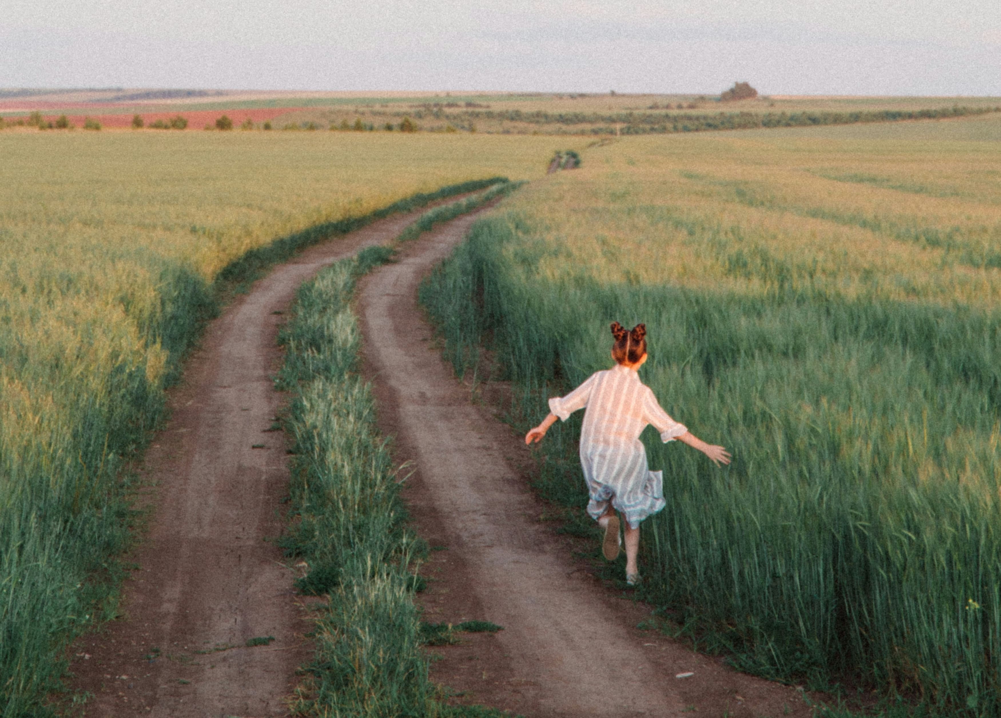 A person in a striped dress runs joyfully along a dirt path through a vast green field under a clear sky.