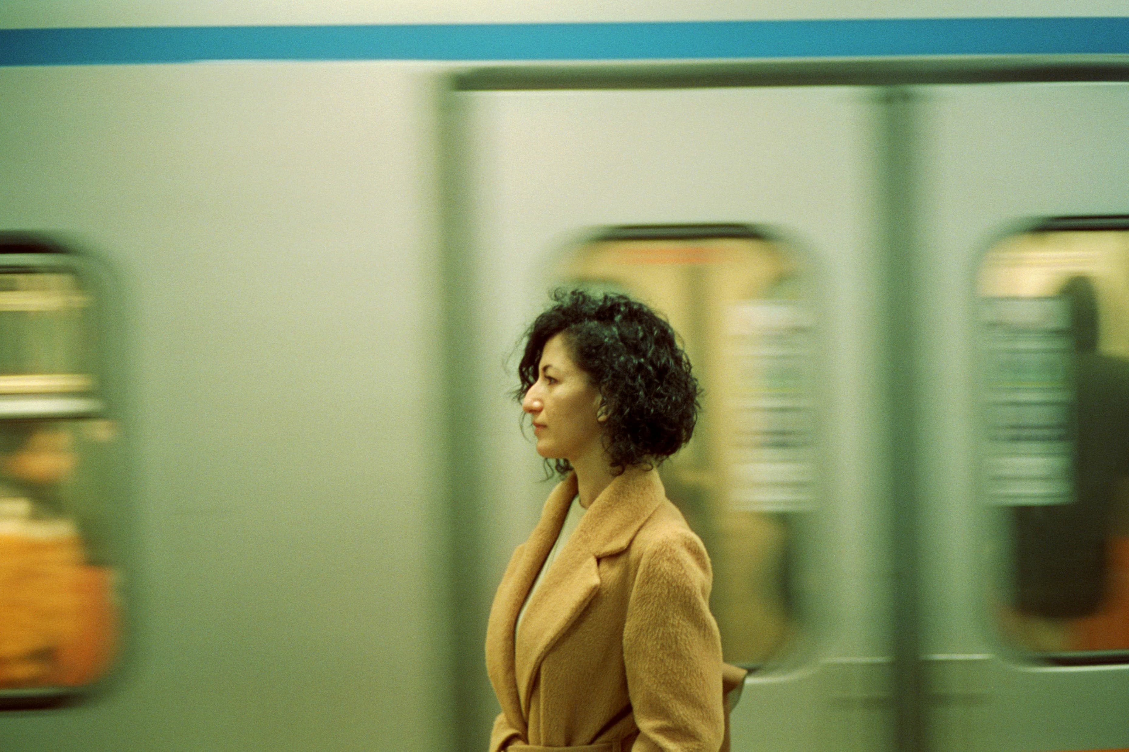 Woman in a brown coat stands still as a subway train moves past her, creating a blurred background effect.