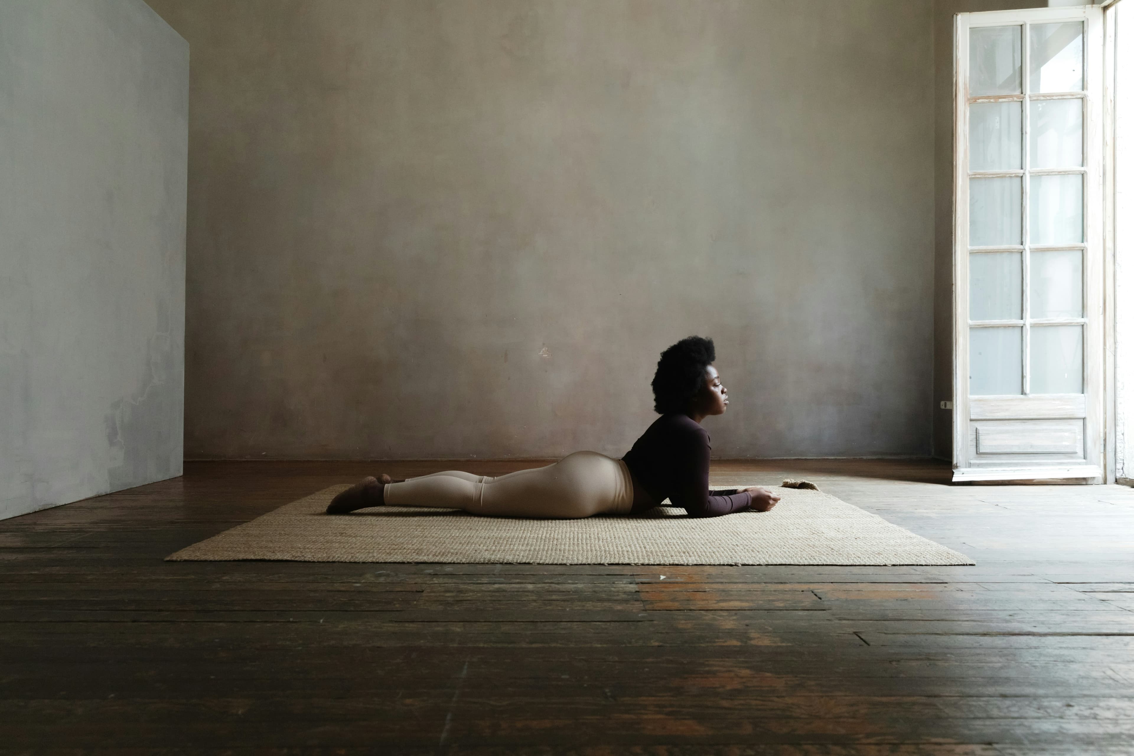 Person in a yoga pose on a mat in a minimalist room with wooden floors and a large window.