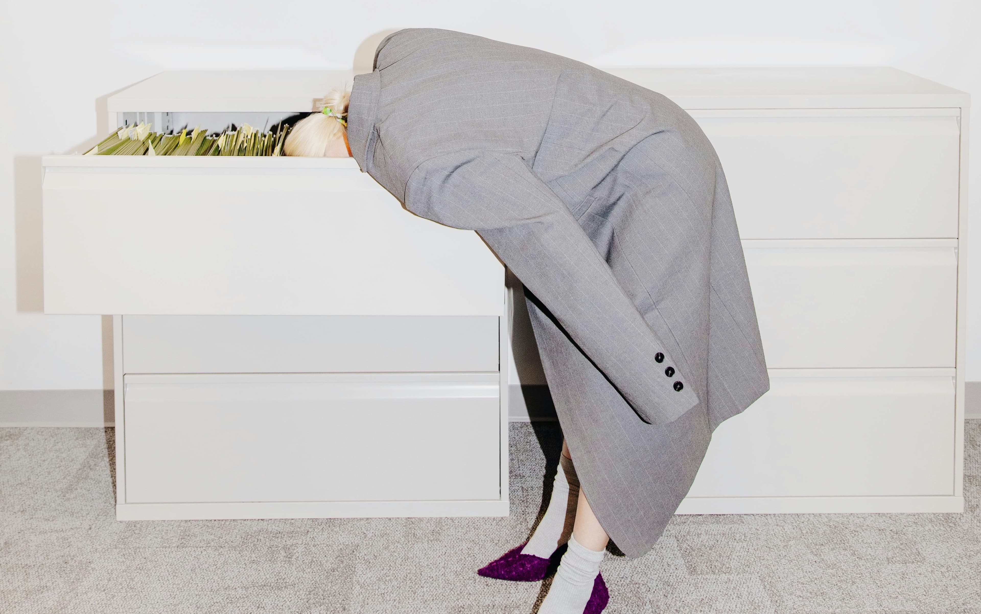 Person in a gray suit with head inside an open filing cabinet drawer, standing in an office setting.