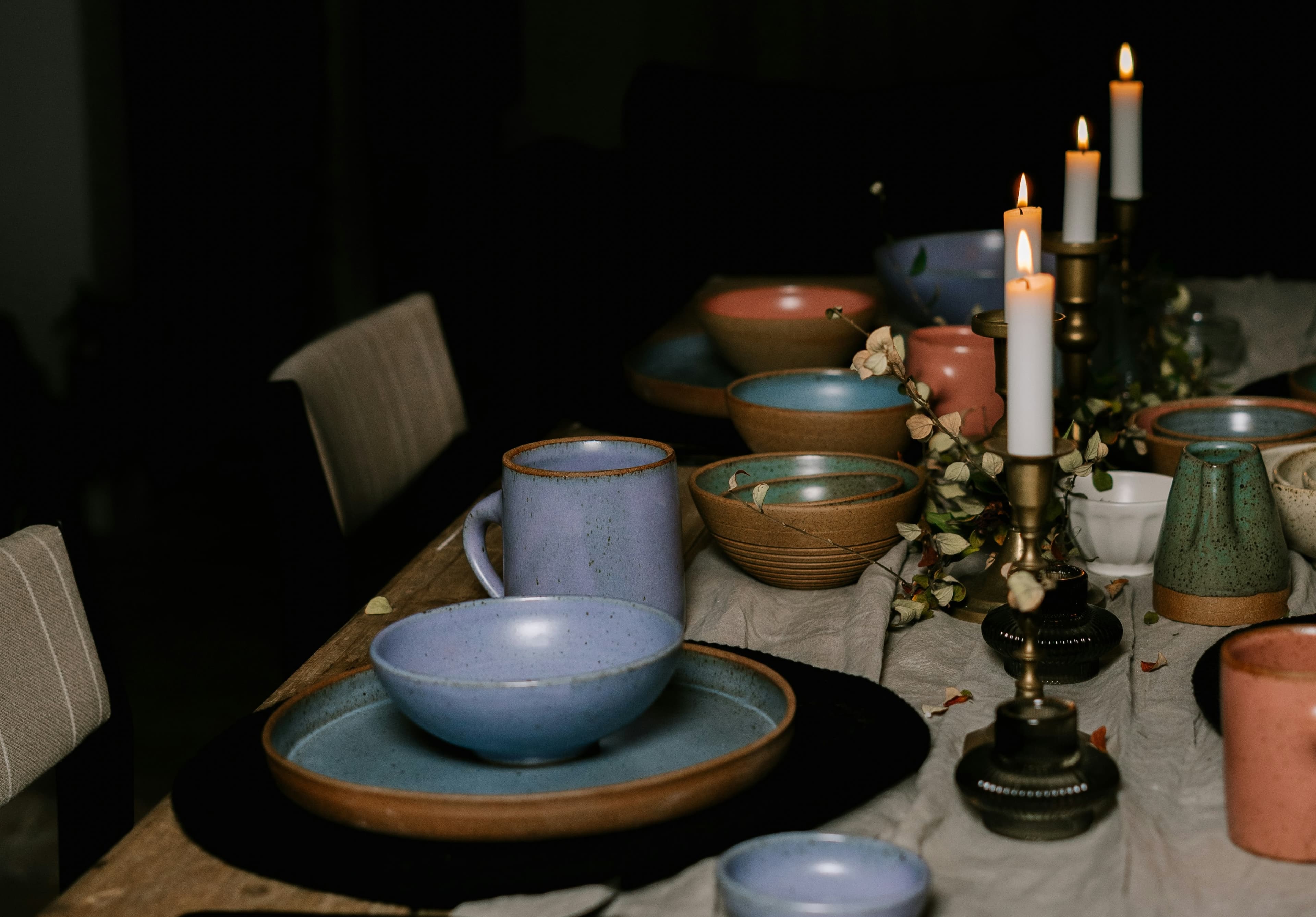A dimly lit dining table with ceramic bowls, mugs, and candles, arranged on a cloth runner, creating a cozy and inviting atmosphere.
