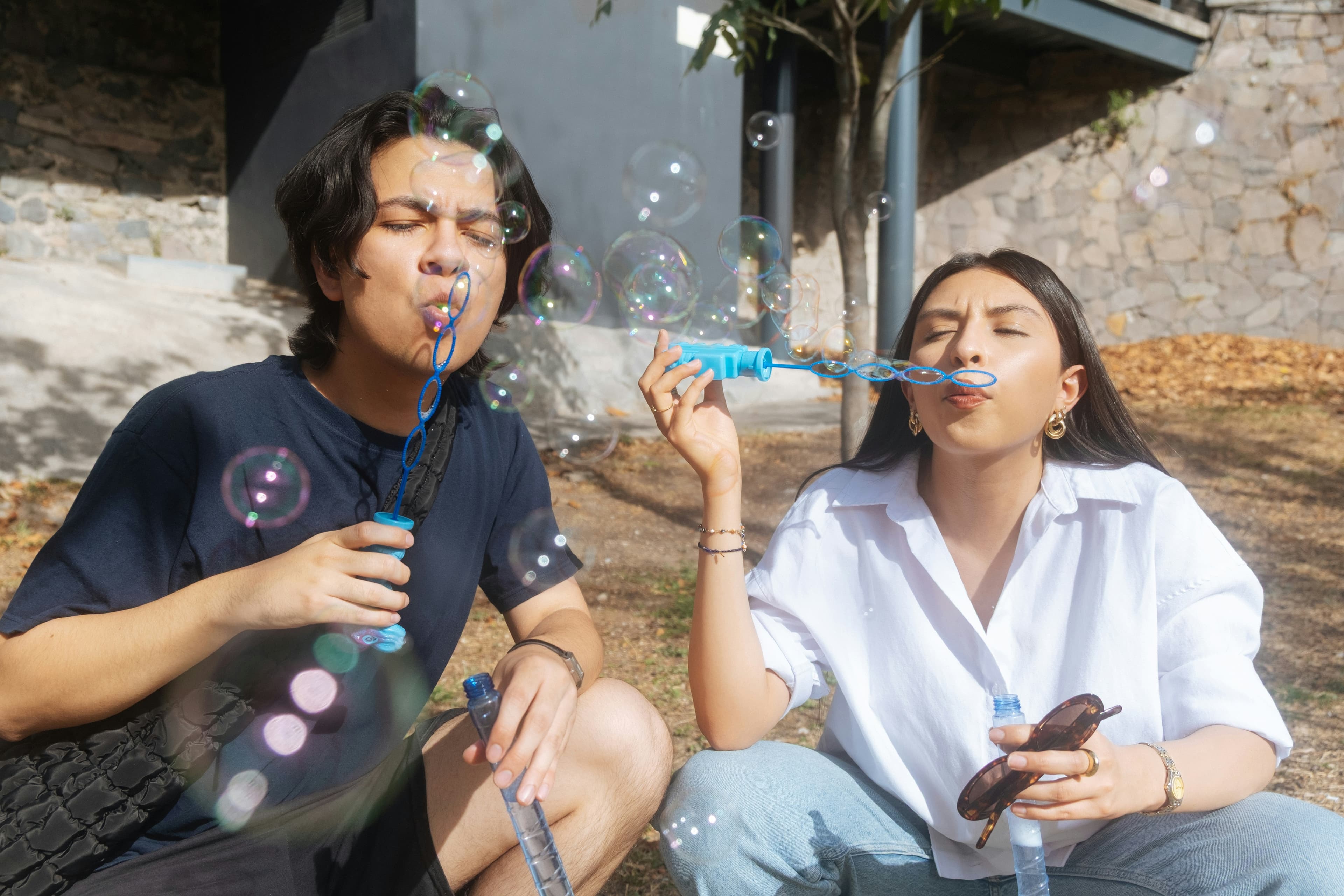Two people sitting outdoors, blowing bubbles with bubble wands. The sun casts shadows, and bubbles float around them.